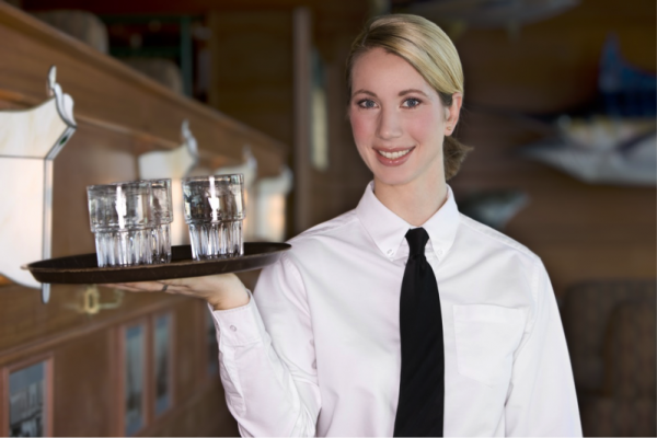 A female server wearing a white shirt and black tie bears a tray of glasses and smiles.
