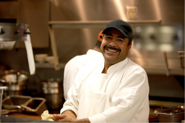 A chef in an industrial kitchen wears a black cap and a wide smile.