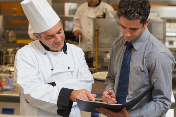 A chef confers with a man in a shirt and tie holding a file. Both men are focused on the file.