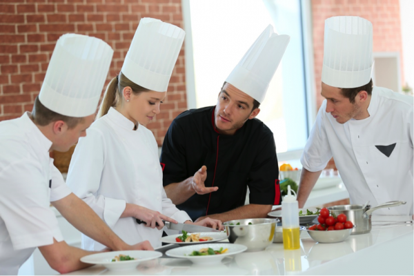 Four chefs in tall white hats prepare a meal together in a room with brick walls and a large window.