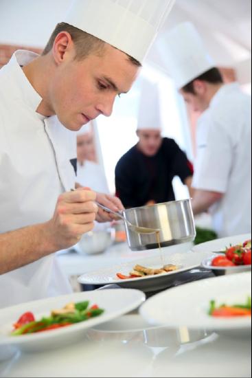 A chef drizzles sauce over a plate. In the background, more chefs work.