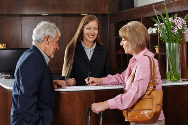 A couple checks into a hotel. Behind the front desk is a smiling woman.