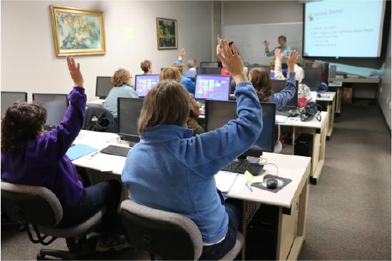 A photograph of a classroom with rows of students sitting behind computers and raising their hands. The instructor stands at the front next to a projected screen.