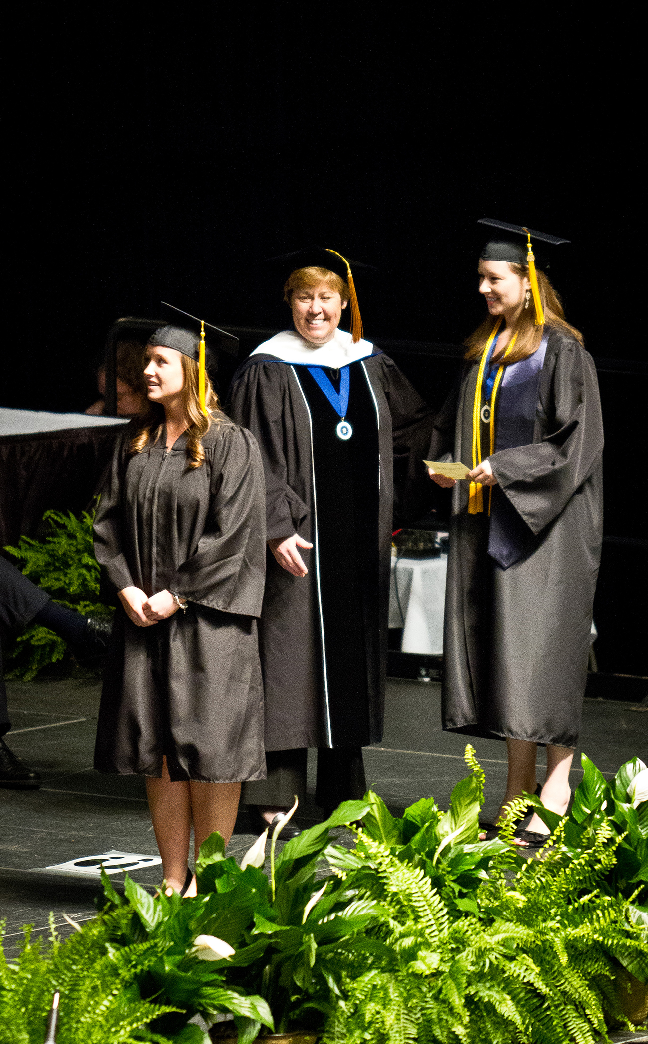 Graduates, wearing cap and gown, walk across a stage to receive their diplomas.