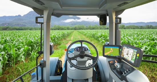 View from the driver's seat of a tractor, looking out over a green field towards mountains and a cloudy sky.