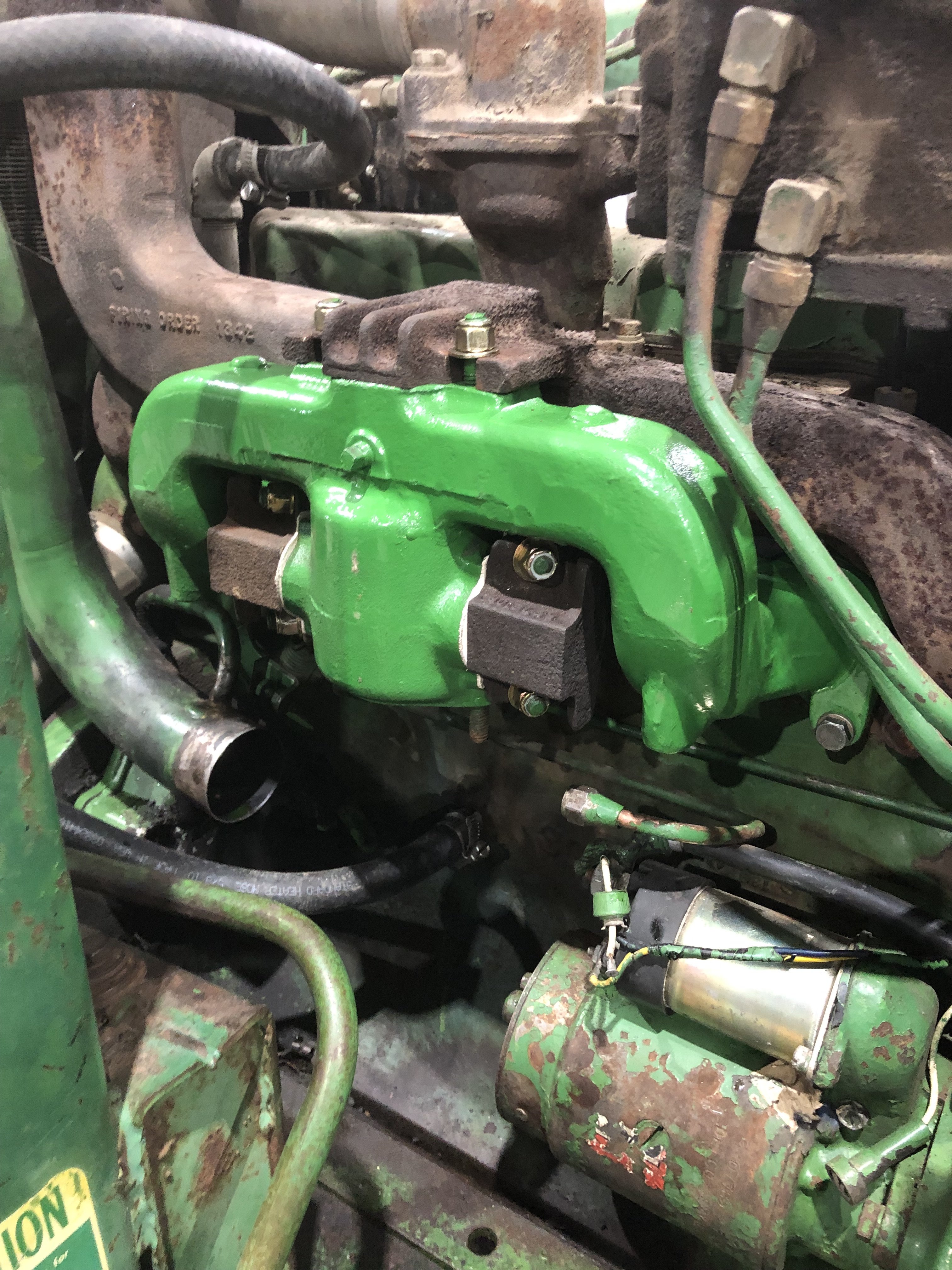 Close-up of a green-painted engine component, showing pipes, bolts, and a starter motor amidst some rust and grime.