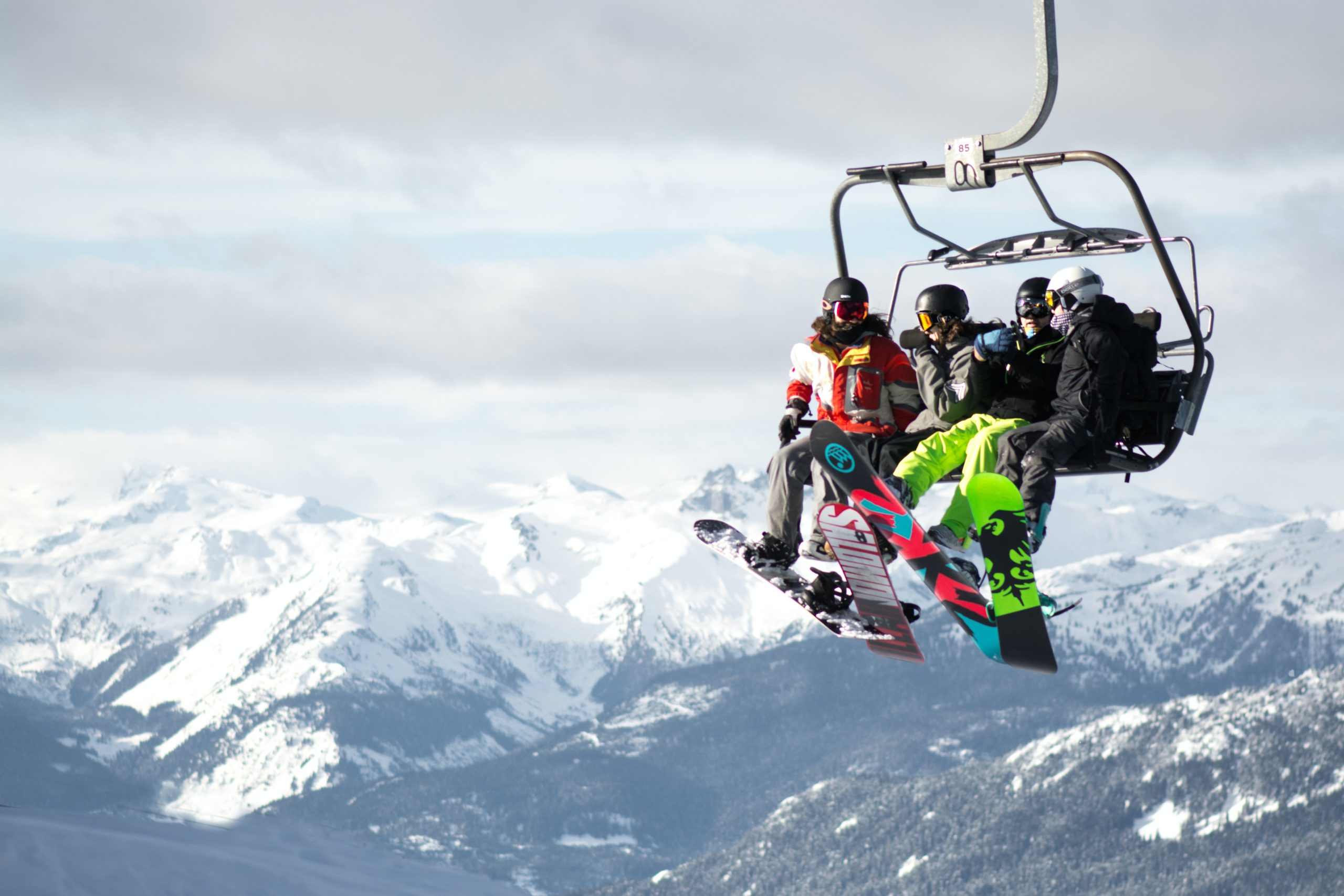 Four snowboarders ride a chairlift high above the ground. Snowy mountains crowd the background.