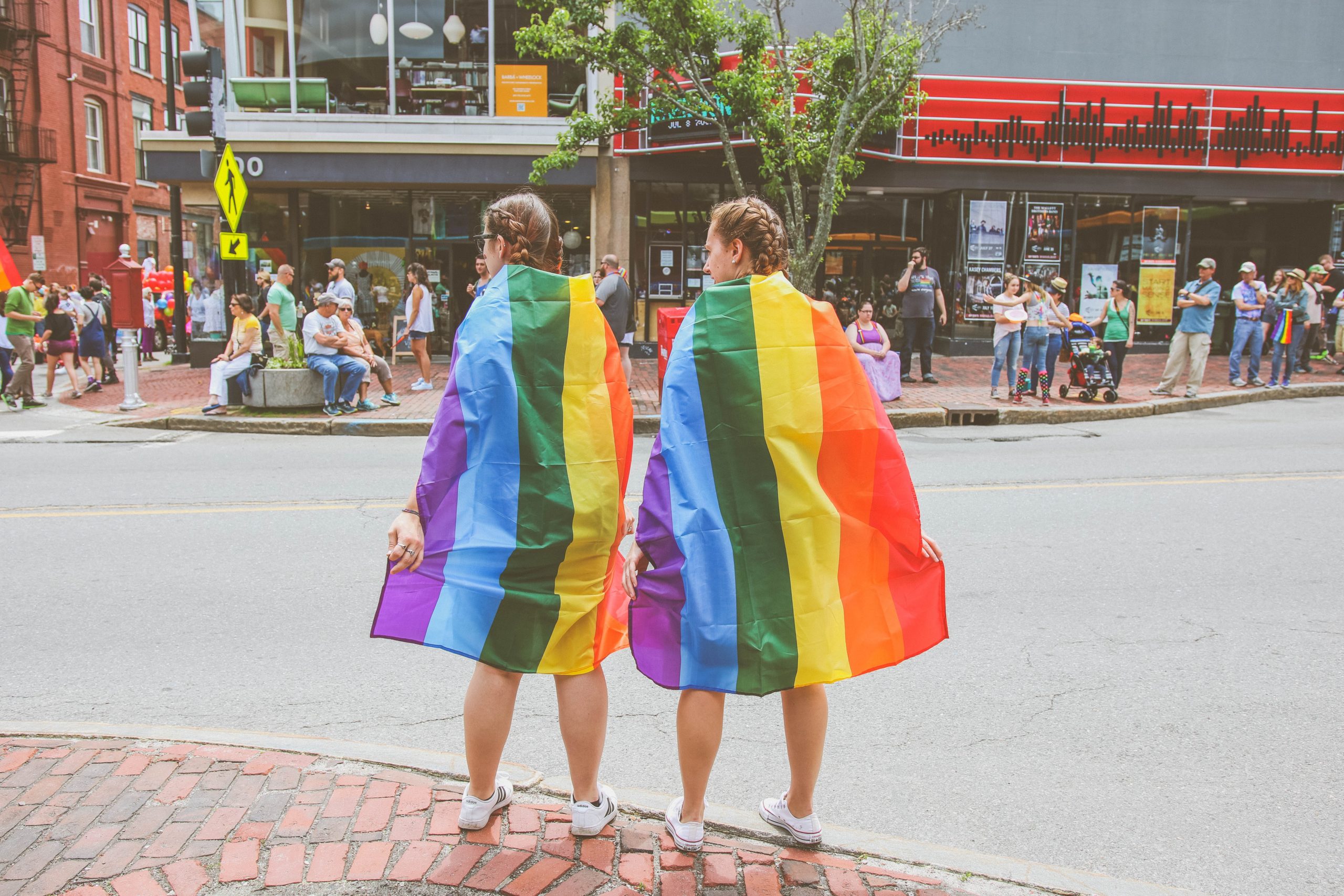 Dos mujeres envueltas en banderas del orgullo arcoíris se enfrentan a una calle de espaldas a la cámara.