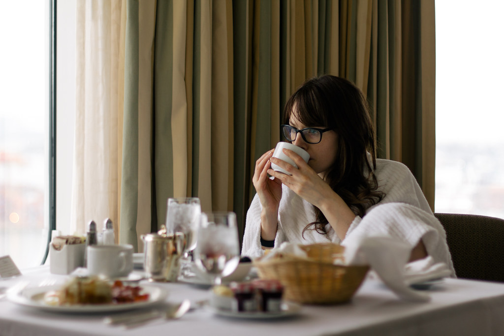 A woman in a bathrobe sips coffee at a small breakfast table next to a huge window.