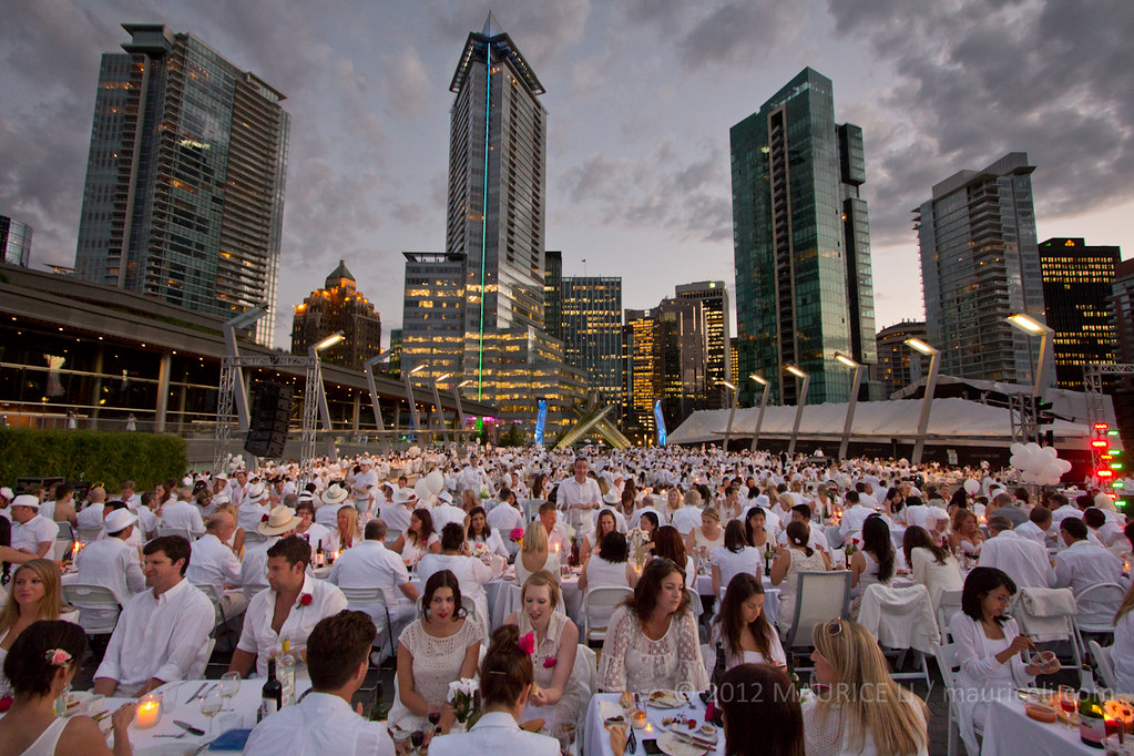Cientos de personas vestidas de blanco cenan al aire libre en mesas pequeñas apiñadas juntas.