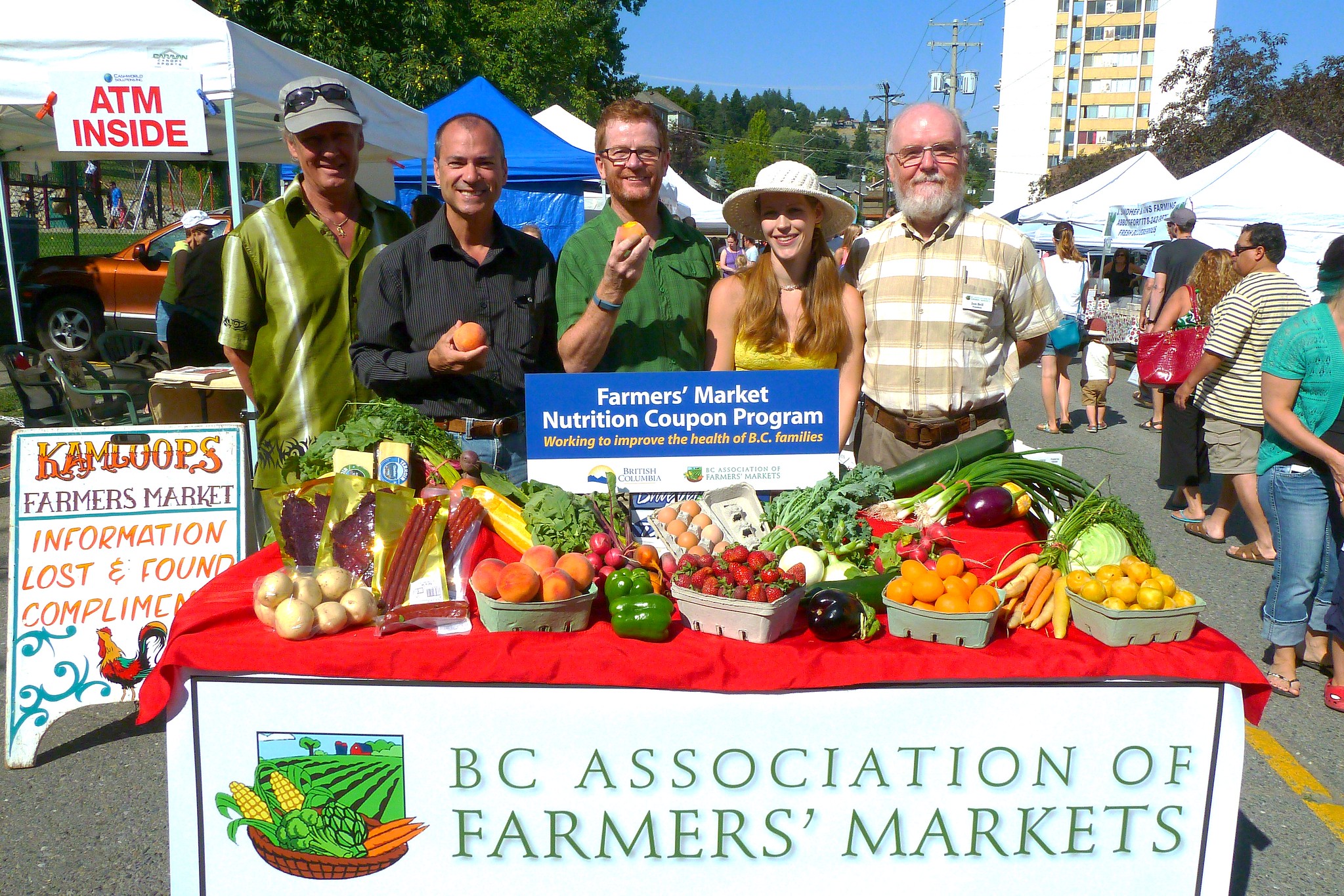 La gente sonríe alrededor de productos frescos y un letrero que dice “BC Association of Farmers' Markets”.
