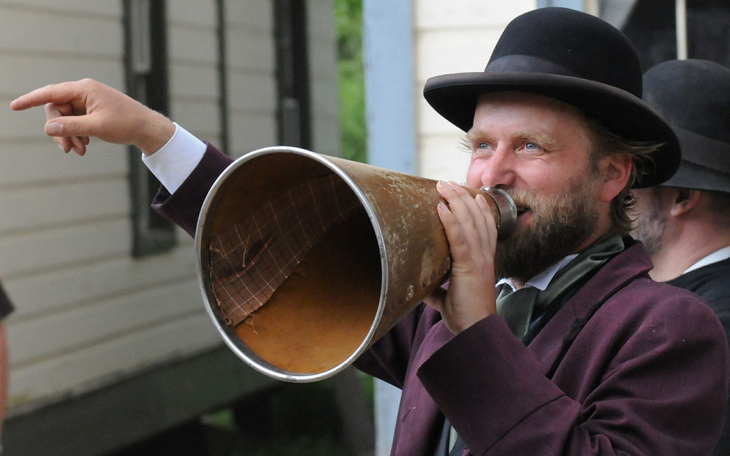 A man dressed in a Victorian suit and bowler hat, speaking into a megaphone.