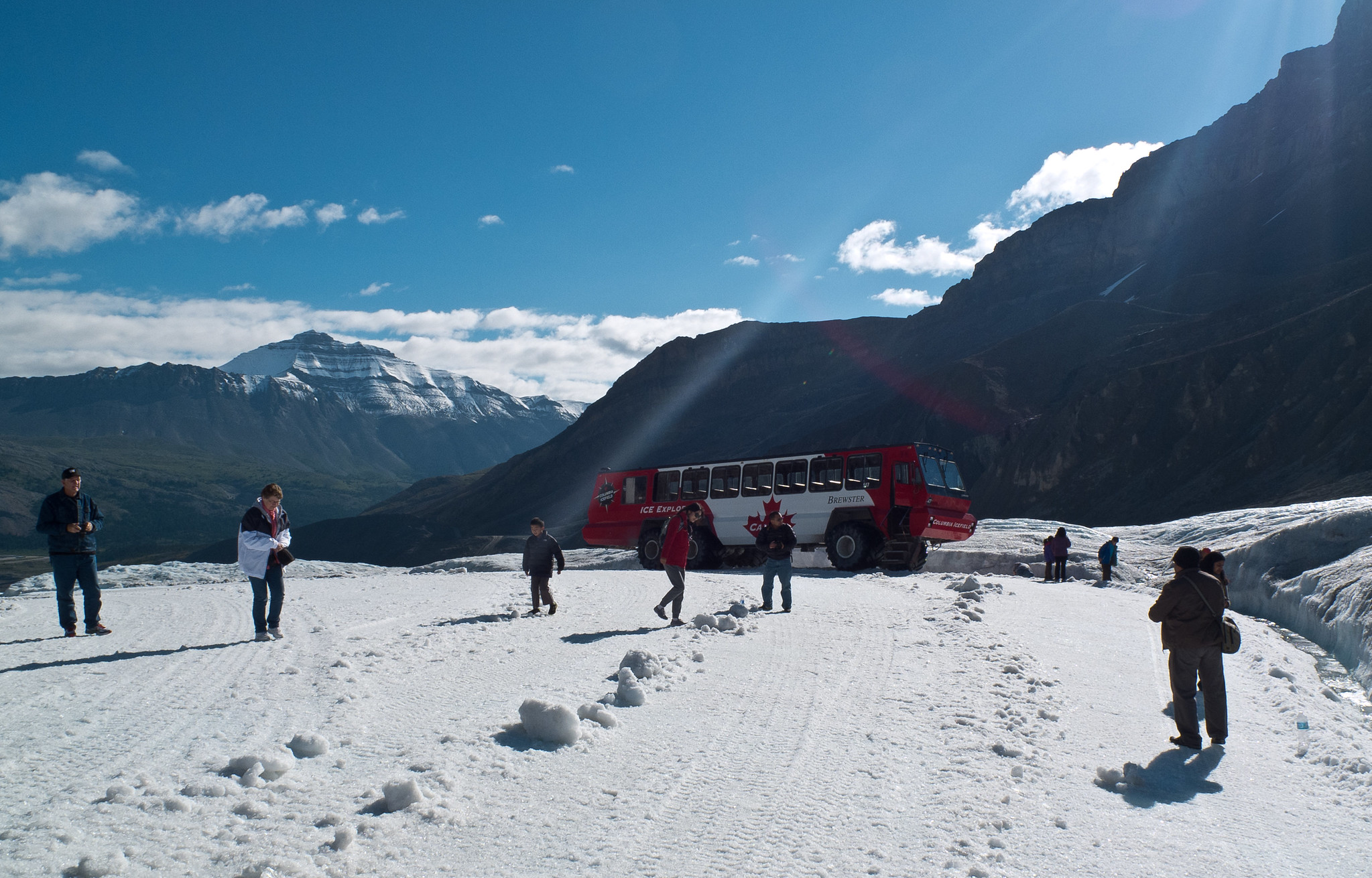 La gente camina por la nieve en las montañas. Un autobús turístico está estacionado detrás de ellos.
