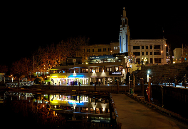 Storefronts by a harbour, lit by streetlamps at night. A clock tower rises from a visitor centre.