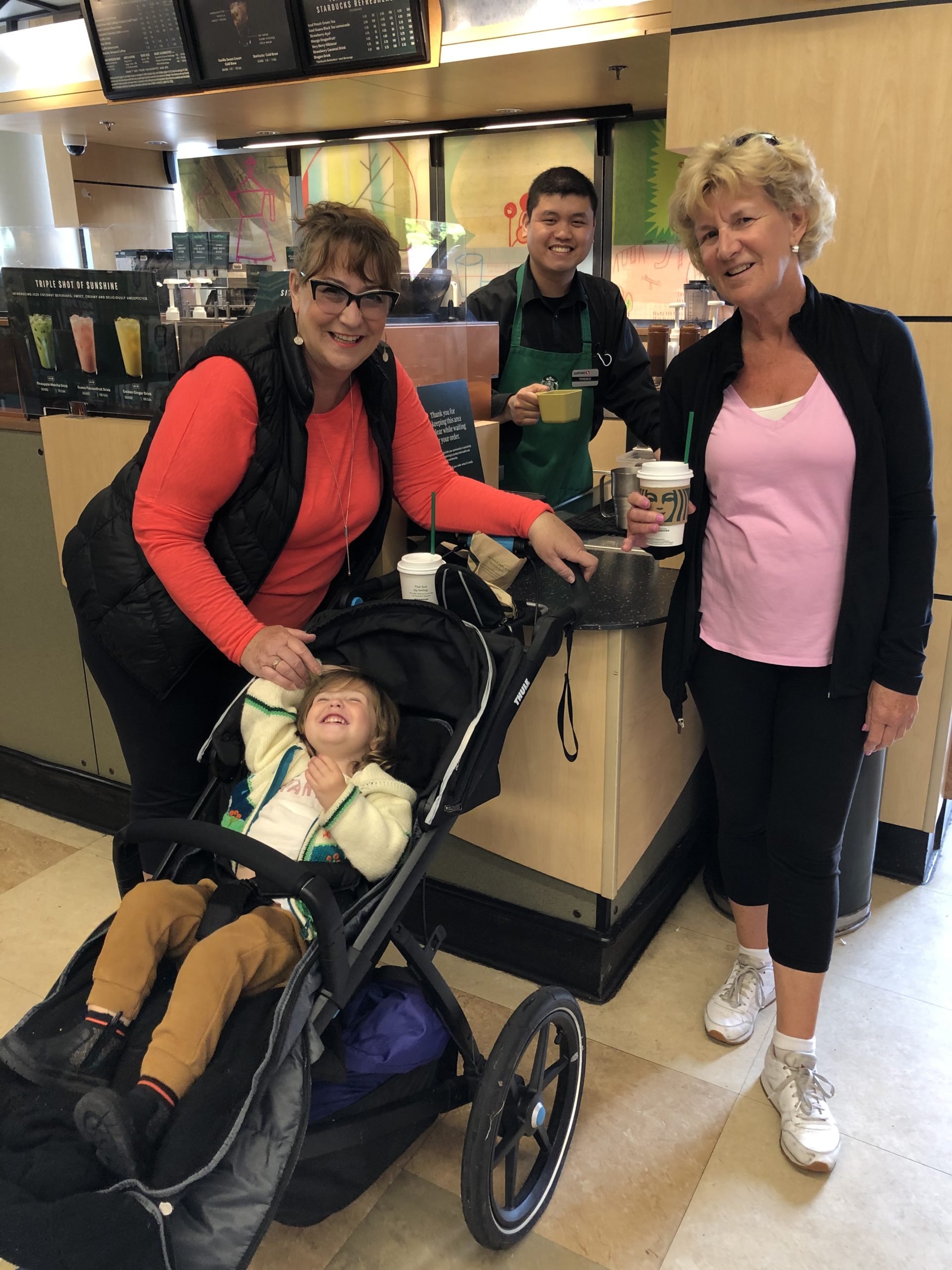 A barista smiles with two customers and a child in a stroller.