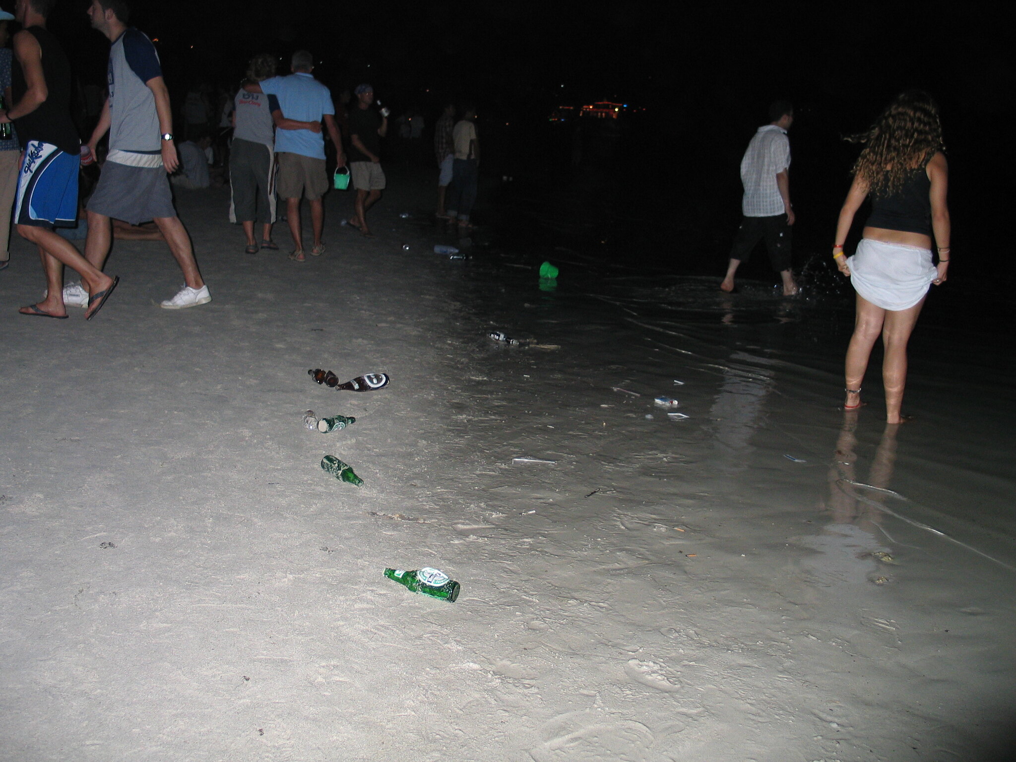 La gente en una playa por la noche le da la espalda a las botellas de cerveza dejadas en la arena.