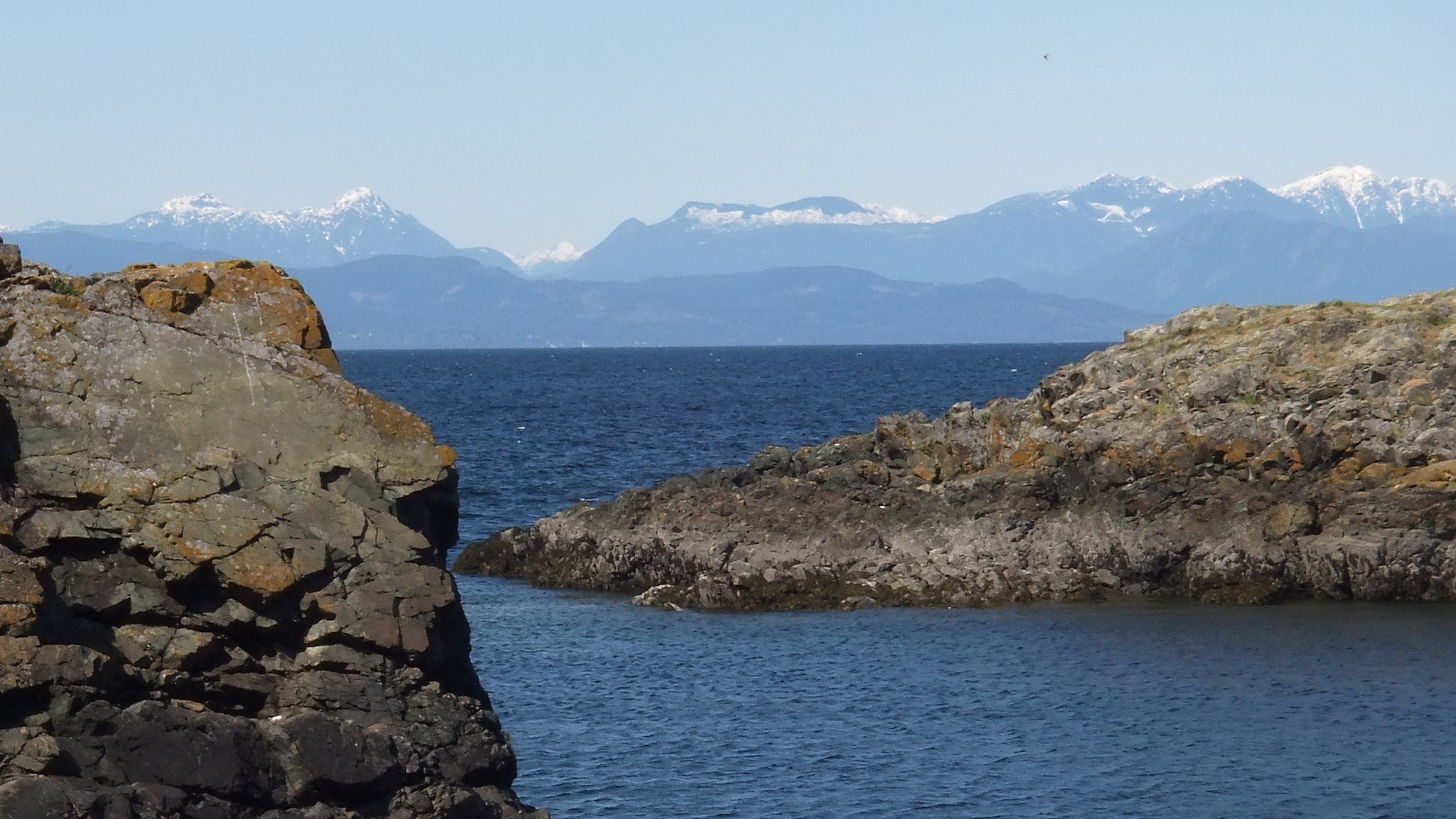 Snowy mountains can be seen across the ocean from a rocky shore.