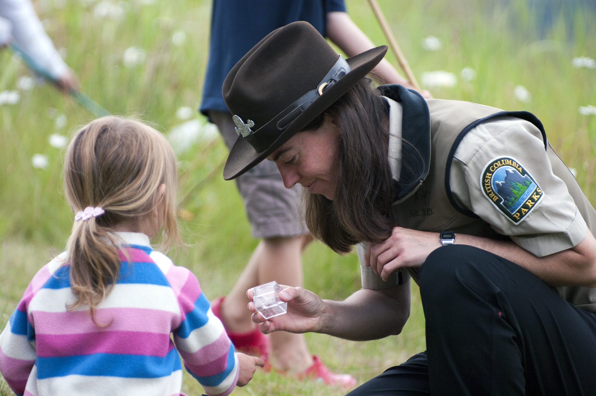 A park ranger holds out a clear plastic cube to a young girl while crouched in tall grass.