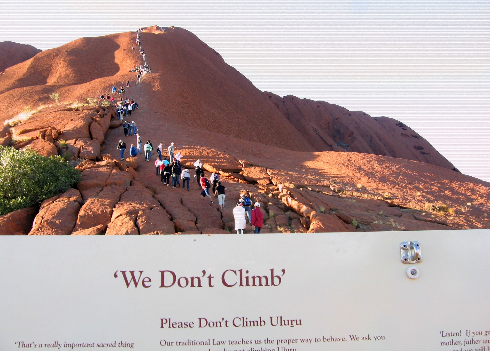 Dozens of people hike over a reddish rock formation.