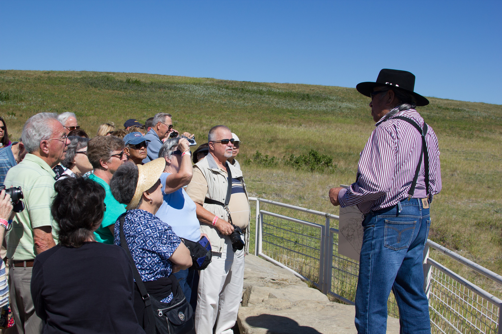 Turistas se reúnen alrededor de un hombre con sombrero vaquero junto a una cerca corta de metal frente a una llanura.
