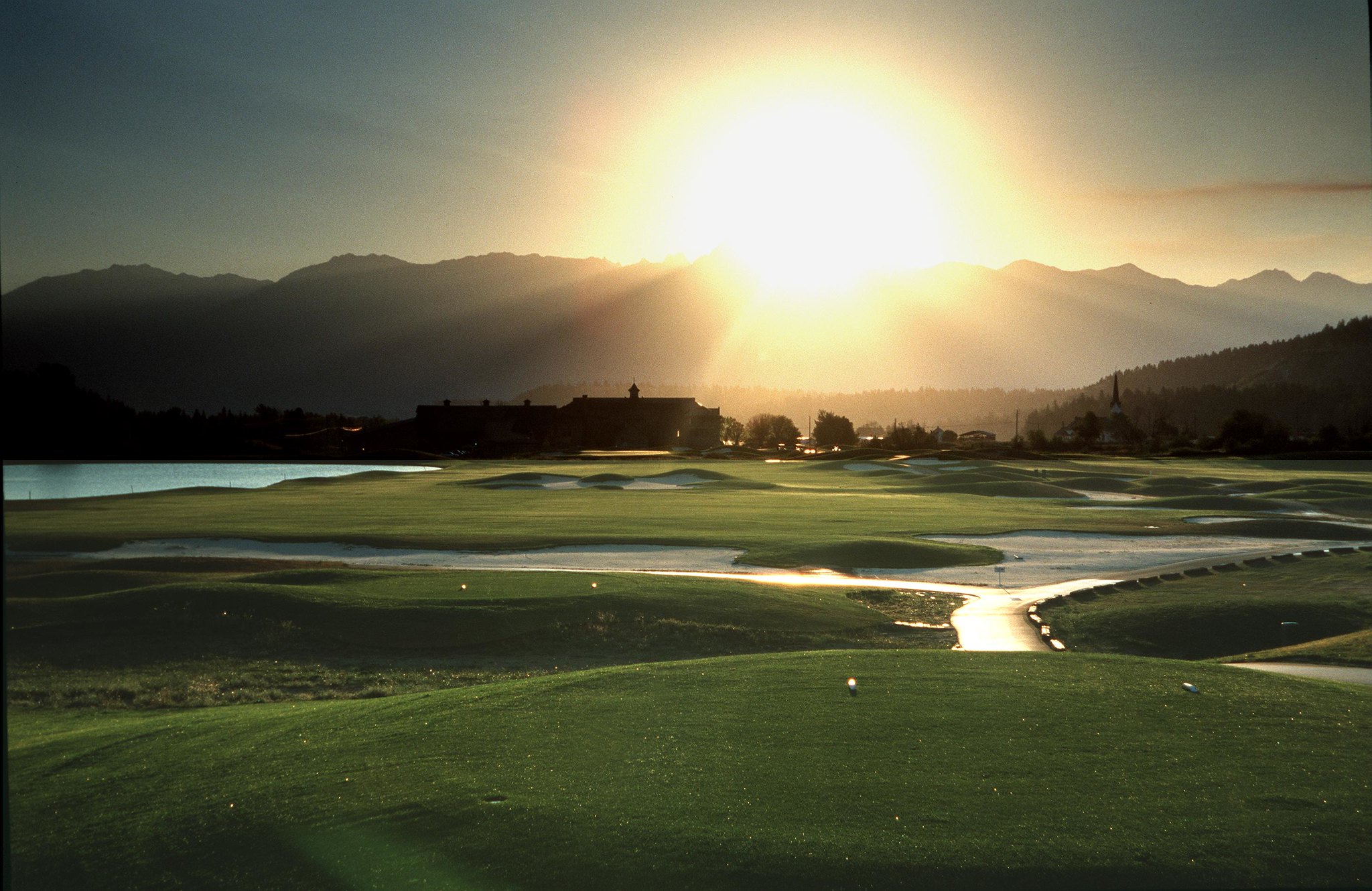 A brilliant sun rises over mountains and shines on a green golf course by the water.