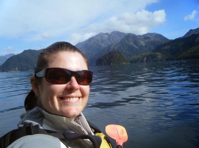 Selfie de una mujer en un chaleco salvavidas sosteniendo una paleta junto a un lago. Las montañas están en el fondo.