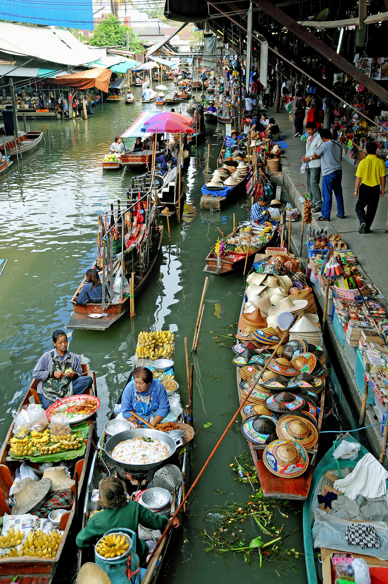 Barcos estrechos llenos de comida, ropa, sombreros y mucho más flotan en un canal junto a un mercado.