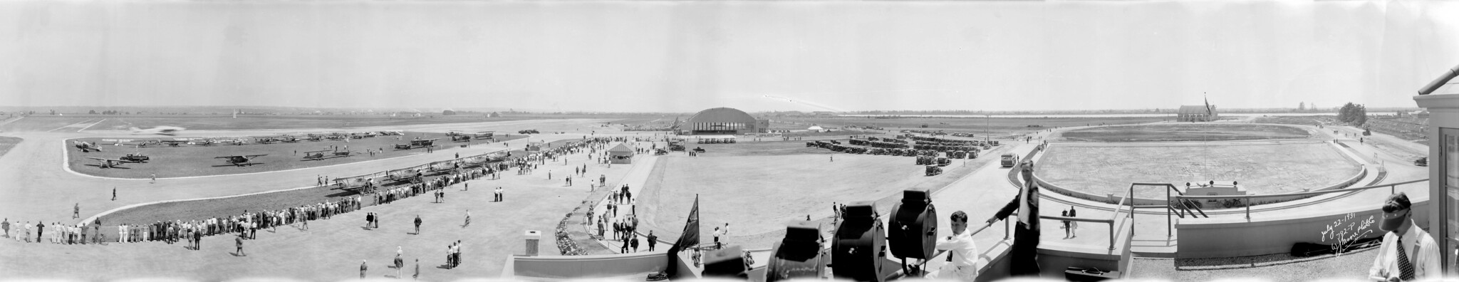 Panorama en blanco y negro de personas alineadas a lo largo de una pista, cerca de varios aviones y un hangar.